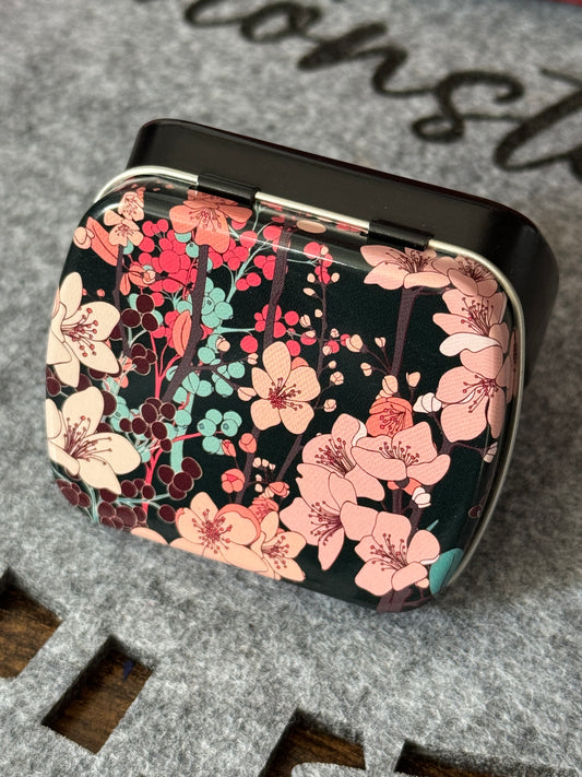 Kleine Weißblechdose, Unterteil schwarz lackiert, Deckel mit floralem Muster in rosa, türkis und braun auf schwarzem Grund.